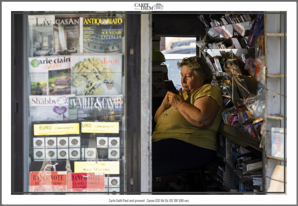 La divisione temporale tra tradizione e innovazione trova espressione e paradosso in questa foto.
Da un lato l’edicola e i suoi colori cartacei, dall’altro l’edicolante che utilizza il proprio device.